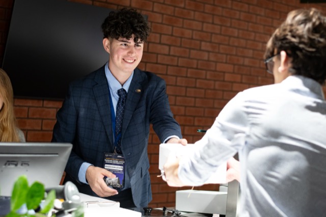 A smiling organizer in a plaid blazer hands a white meal box to a delegate across the counter.