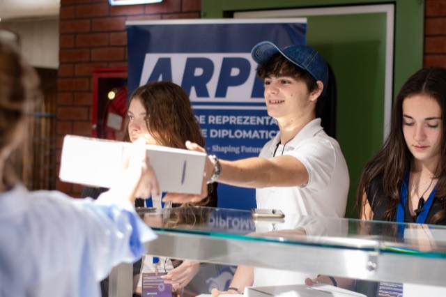A volunteer in a denim cap passes a boxed meal over the glass counter beside the ARPD banner.