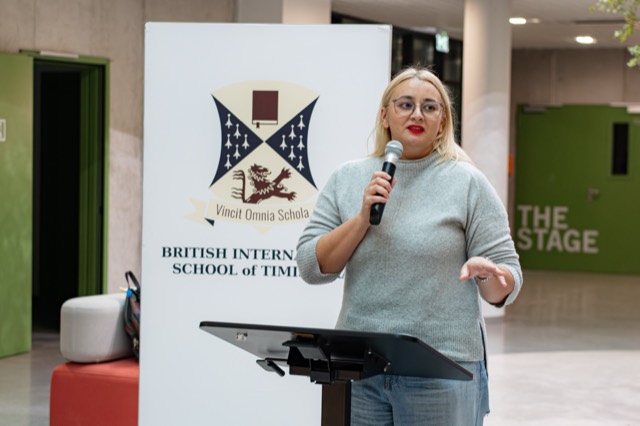 A speaker with a microphone addresses the ceremony beside the school banner.