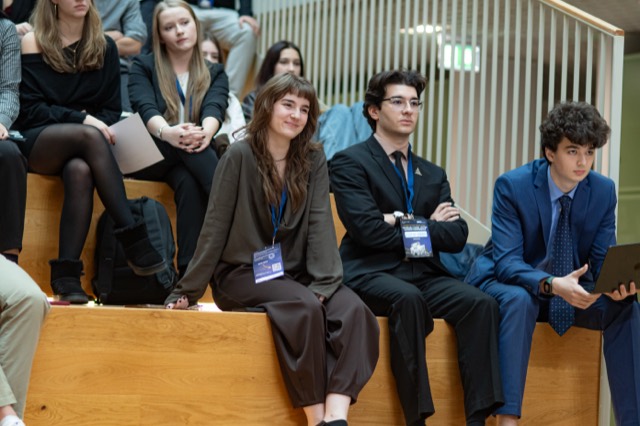 Three delegates sit on a tiered bench, one balancing a laptop on his knee.